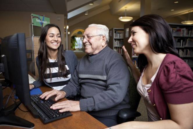 Foto oudere man achter een computer met 2 jonge vrouwen die hulp bieden